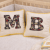 Pair of personalized 25x25cm children cushions with African print appliqué initials, beige fabric, contrast piping, styled in a cozy nursery.