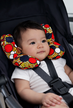 Newborn baby seated in a car seat wearing an AfriBloom wax fabric support pillow, shown in everyday travel setting