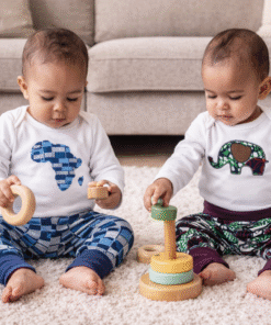 Two babies wearing MiniBloom African appliqué baby outfits, playing on a living room carpet