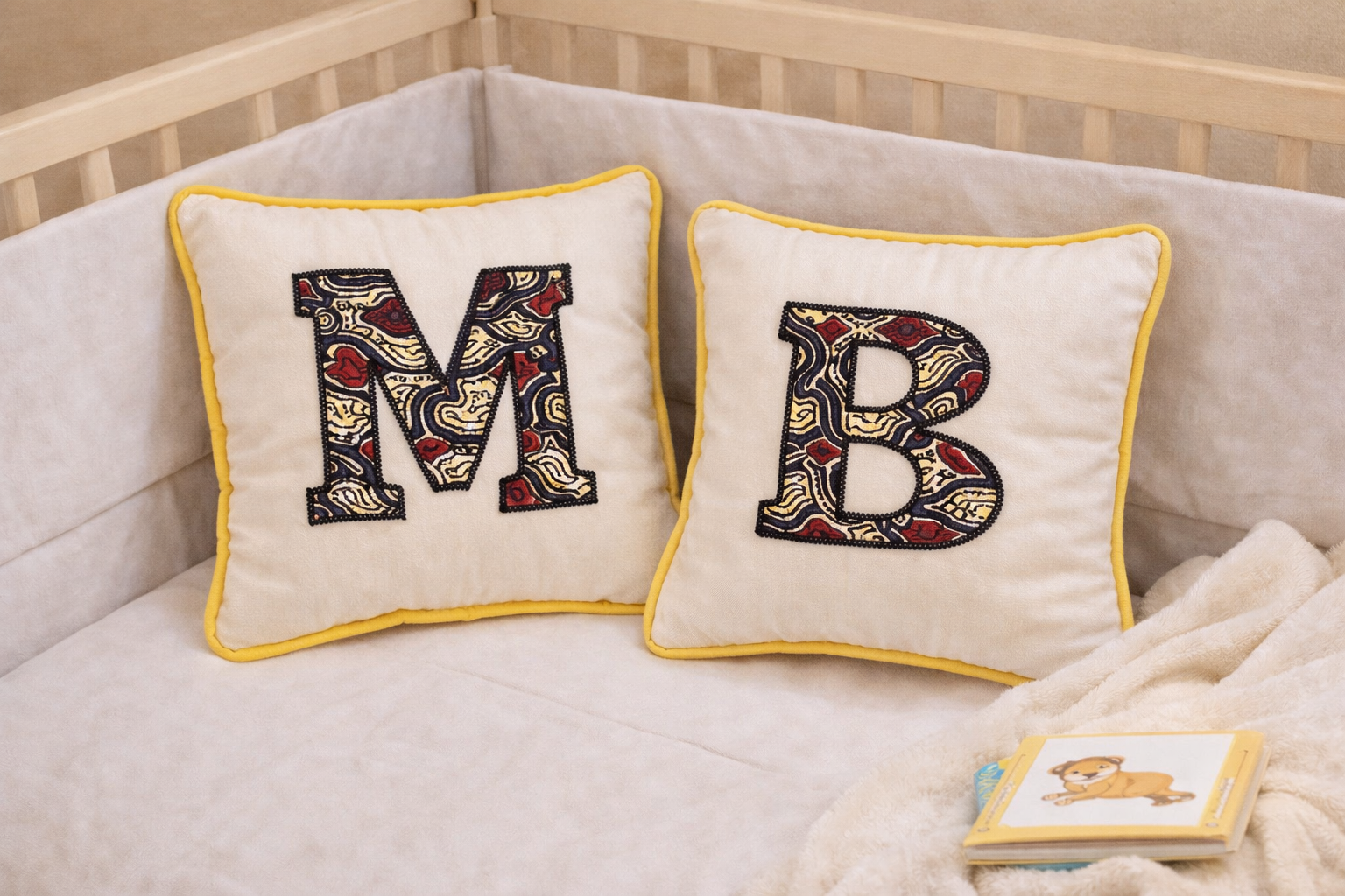 Pair of personalized baby initial cushions with African print appliqué letters placed in nursery crib, handmade decorative pillows for baby room