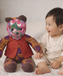 AfriBloom handmade baby bear plush toy with African wax fabric, shown next to a smiling baby in a bedroom